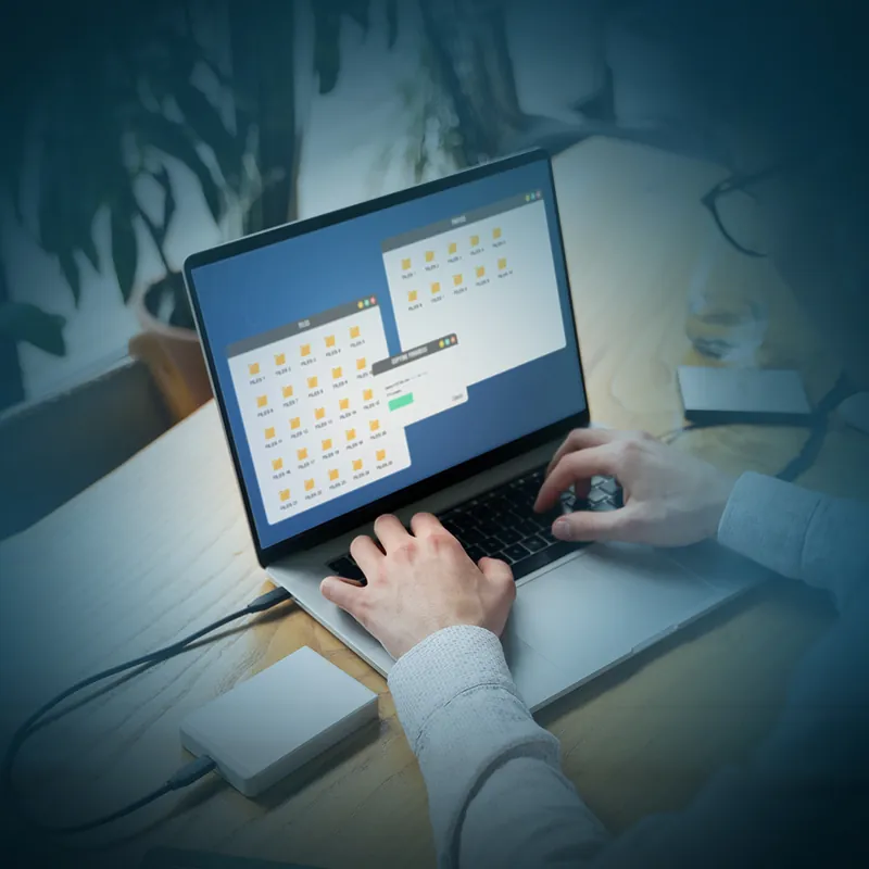 Two hands typing on a laptop whose screen shows folder windows and an operation in progress, with an external drive on the desk